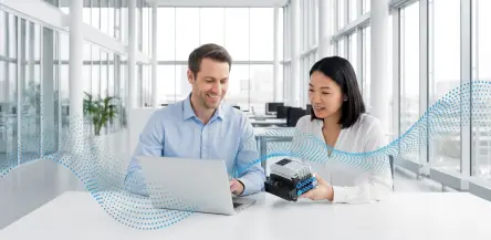 A man and a woman sit at a desk in an office and use the Festo Virtual Assistant to quickly look up product data for the valve terminal she holds in her hand.