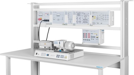 Imagen de un banco de trabajo industrial o laboratorio de formación en electrotecnia, con un motor eléctrico montado sobre una mesa y conectado a un módulo de control. Al fondo se observan paneles didácticos con esquemas eléctricos y diagramas, además de equipos de medición y control dispuestos en una estructura tipo rack.