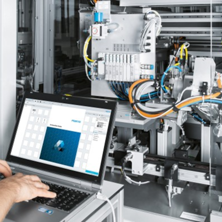 A technician operates a laptop connected to an automated industrial machine, monitoring system data and controls in a manufacturing environment.