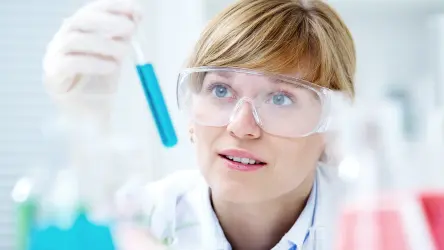 A imagem mostra uma cientista mulher em um laboratório, examinando cuidadosamente um tubo de ensaio com um líquido azul. Ela usa óculos de proteção e uma bata de laboratório branca. O ambiente ao redor é iluminado e parece ser um laboratório bem equipado com diversos frascos e equipamentos de laboratório. A expressão dela é focada e curiosa enquanto ela observa a amostra.