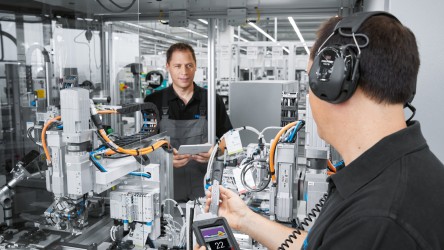 A imagem mostra dois técnicos em um ambiente industrial, provavelmente dentro de uma fábrica ou unidade fabril. Um técnico está em primeiro plano, usando um dispositivo portátil, possivelmente para diagnóstico ou controle de máquinas. Ele usa um fone de ouvido, sugerindo que pode estar conduzindo uma operação guiada ou consulta remota. O outro técnico, ao fundo, observa ou monitora máquinas, segurando um tablet que pode exibir dados operacionais ou controles. O ambiente está repleto de máquinas complexas, indicando um chão de fábrica de alta tecnologia.