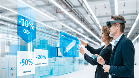 A imagem mostra dois profissionais, um homem e uma mulher, usando dispositivos de realidade aumentada em uma fábrica. Eles estão interagindo com gráficos digitais holográficos que flutuam no ar à frente deles, exibindo estatísticas como 