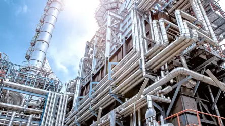 A imagem mostra uma planta industrial robusta sob um céu claro. A estrutura é complexa, com muitas tubulações de metal prateadas, colunas e escadas de segurança. Há também chaminés altas e várias seções modulares que indicam uma operação de grande escala. A luz do sol ilumina a instalação, realçando os detalhes metálicos e criando um contraste vibrante com o céu azul ao fundo.