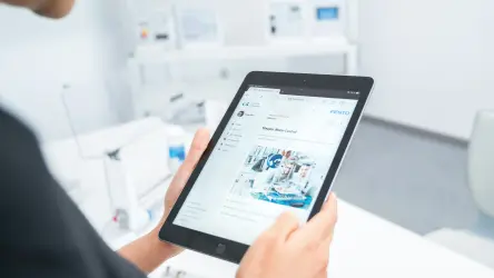 A imagem mostra uma pessoa, visível da cintura para cima, segurando um tablet e navegando em um site com informações sobre controle de motores de passo. O tablet exibe uma página da Festo, uma empresa conhecida por suas soluções de automação. O indivíduo está em um ambiente de laboratório ou sala de aula técnica, com equipamentos e painéis de controle ao fundo, sugerindo um cenário de treinamento ou trabalho em automação industria
